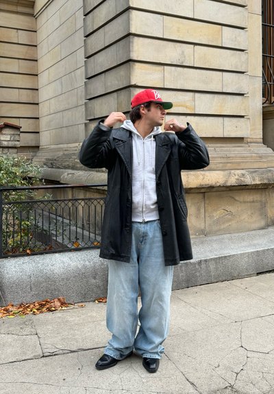 Abrigo de cuero negro sobre una sudadera gris, combinado con jeans ligeros y sueltos de color azul claro y zapatos negros. Gorra roja usada al revés, de pie al aire libre.