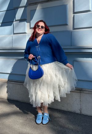 Cárdigan azul de punto con cierre de lazo, falda de tul blanca en capa, bolso azul y zapatillas de deporte azul claro, todo contra un fondo de pared azul.
