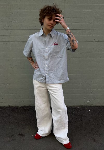 Camisa de botones a rayas en azul claro y blanco con bordado en rojo, combinada con pantalones anchos blancos y calcetines rojos, mostrando un ajuste relajado.