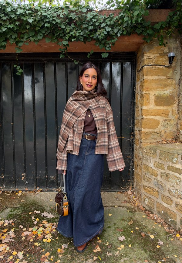 Une femme pose en plein air, vêtue d’une veste-cape à carreaux, d’une longue jupe en jean et de bottes brunes. Elle tient un petit sac à main près d’un mur de pierre et d’une porte noire.