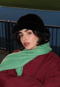 Μαύρο καπέλο τύπου bucket από γούνα, πράσινο υφασμάτινο κασκόλ και μπορντό φουσκωτό μπουφάν. Το υπόβαθρο περιλαμβάνει μια μπλε πίστα δρομολόγησης και μια μαύρη περίφραξη.