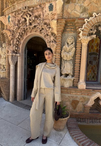 Mujer con conjunto beige y zapatos oscuros, de pie junto a un arco de ladrillo ornamentado y una estatua de piedra cerca de una ventana de vidrieras y una planta en maceta.