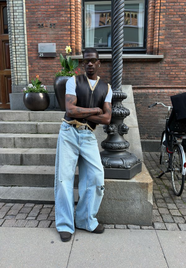Uomo con occhiali da sole, giacca senza maniche e jeans larghi appoggiato a un ornato palo di lampada nero sui gradini del marciapiede cittadino.