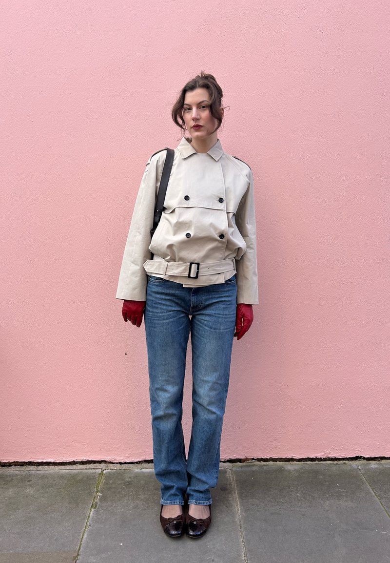 Femme portant une veste beige ceinturée, un jean bleu, des gants rouges et des chaussures marron, debout contre un mur rose avec un sac à bandoulière noir.