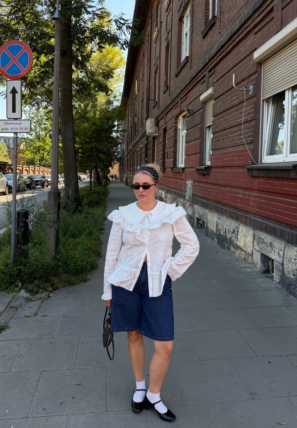 Kvinde iført hvid bluse med flæser, denimshorts, sorte sko med hvide sokker og solbriller står på fortovet ved siden af en murstensbygning.