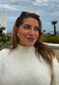 Λευκό πουλόβερ με ζιβάγκο σε πλέξη, που διαθέτει μια υφαντή διαμαντένια υφή. Μεγάλοι χρυσοί κρίκοι στα αυτιά και φόντο με συννεφιασμένο ουρανό και φοίνικες.