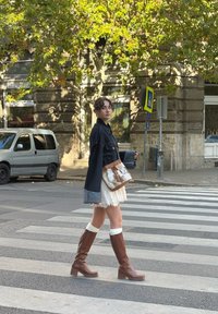 Casaco de ganga com mangas contrastantes, vestido de renda, botas castanhas de couro até aos joelhos com tacões bloco, e uma mala texturizada com franjas, numa passadeira.