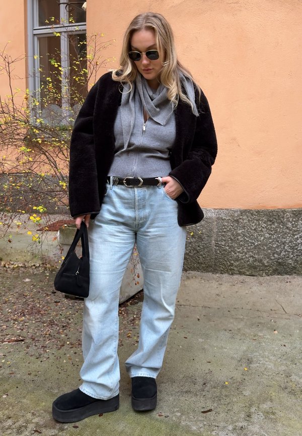 Kvinne iført solbriller, svart jakke, grå hettejakke med glidelås, lyseblå jeans og svarte platåsko, holder en svart håndveske utendørs.