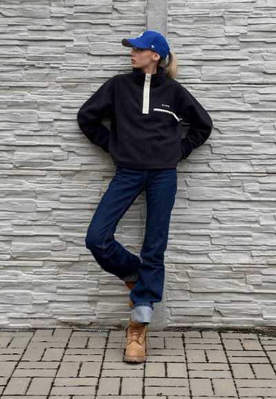 Sudadera de forro polar negra con un cuello alto y botones crema, combinada con jeans de campana azul oscuro y botas marrón claro. Una gorra azul completa el conjunto.