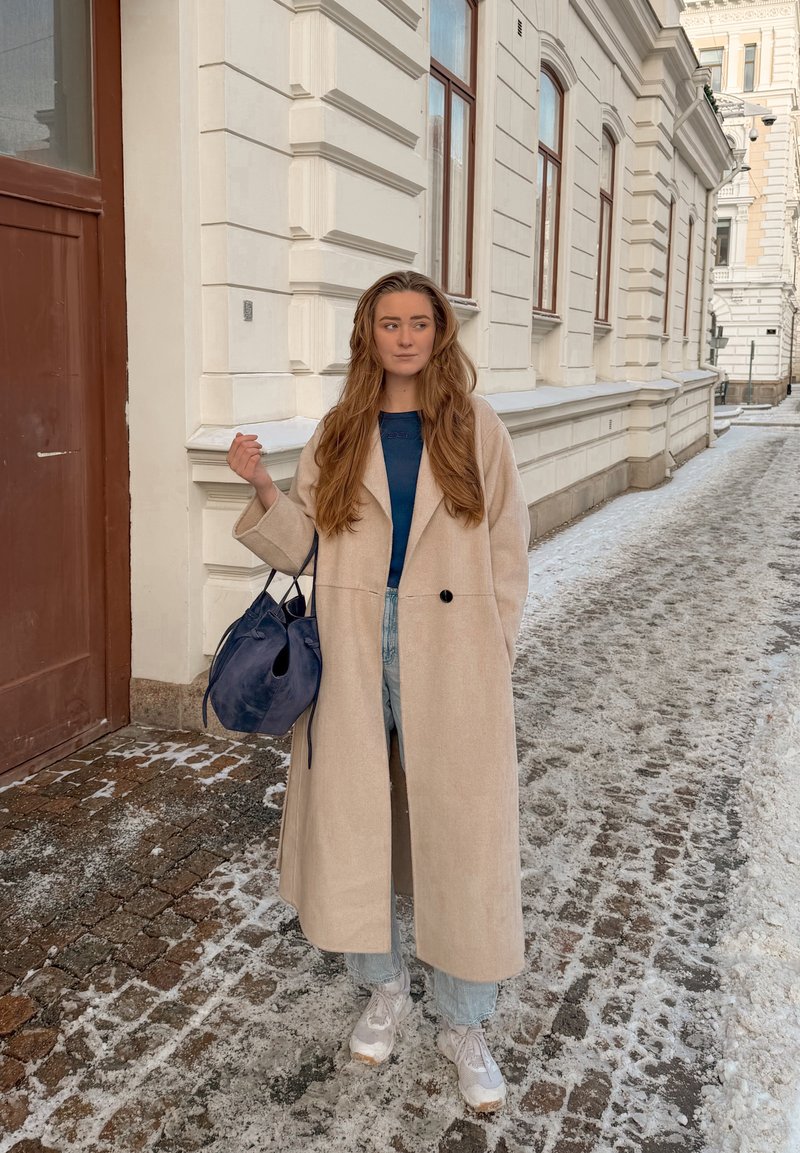 Femme en long manteau beige, haut bleu, jean clair et baskets blanches, tenant un sac à main bleu marine, debout sur une rue pavée enneigée près d'un bâtiment.