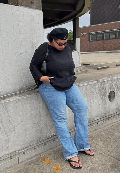 Camisa negra de manga larga, jeans azul claro con botamanga acampanada y sandalias negras. Los accesorios incluyen un pequeño bolso negro y gafas de sol.