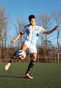 Jeune joueur de football masculin en tenue de l'Argentine, en train de frapper un ballon sur un terrain vert, avec des arbres sans feuilles et un ciel clair en arrière-plan.