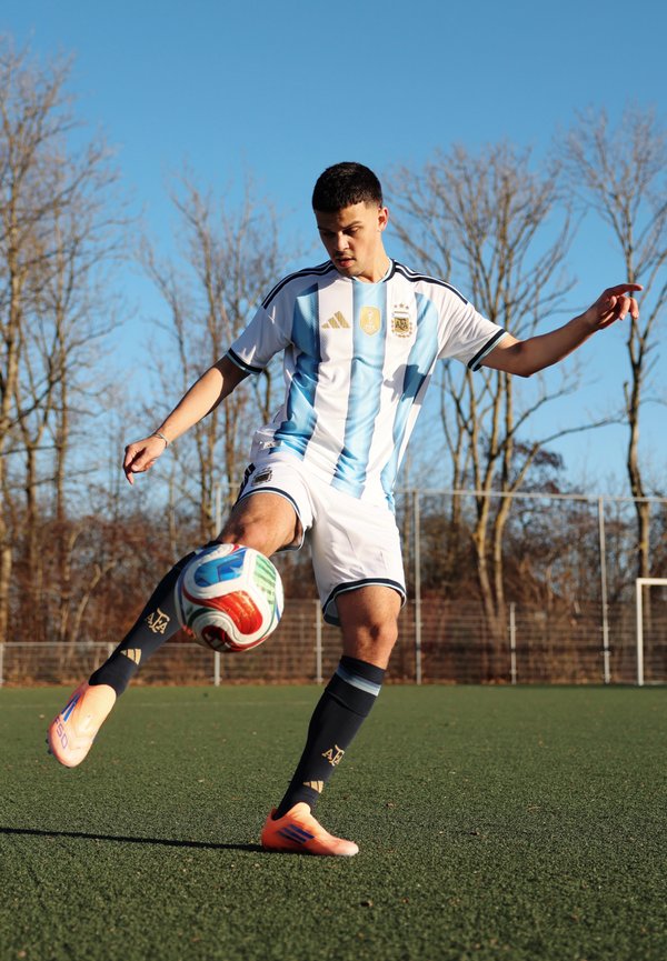 Jeune joueur de football masculin en tenue de l'Argentine, en train de frapper un ballon sur un terrain vert, avec des arbres sans feuilles et un ciel clair en arrière-plan.