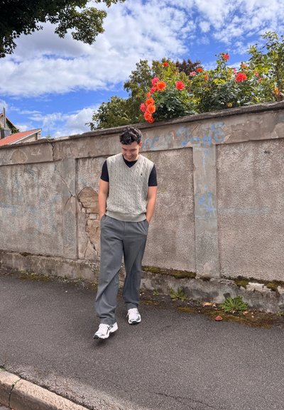 Chaleco de punto gris sobre una camiseta negra, combinado con pantalones grises y zapatillas blancas, con un fondo de una pared de piedra texturizada y flores.