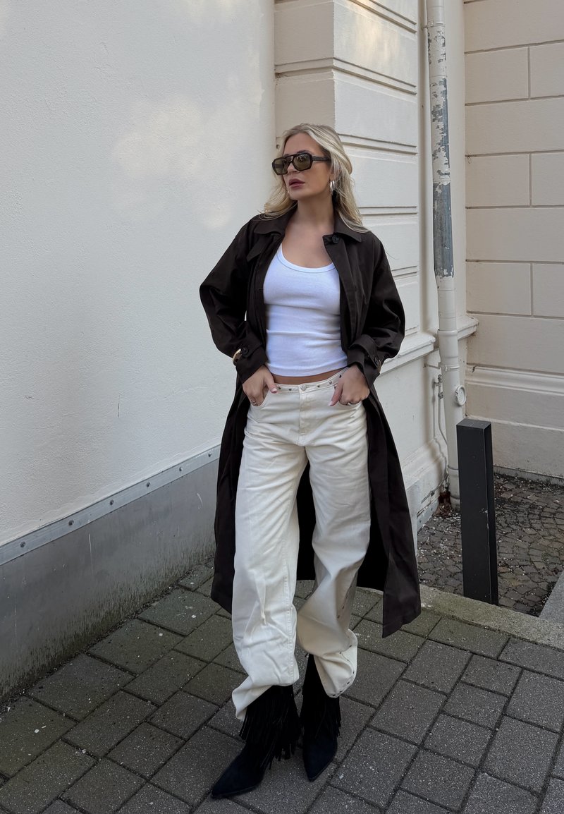 Femme portant un manteau long foncé, un débardeur blanc, un pantalon crème, des bottes noires et des lunettes de soleil, debout sur un trottoir pavé près du coin d'un bâtiment.