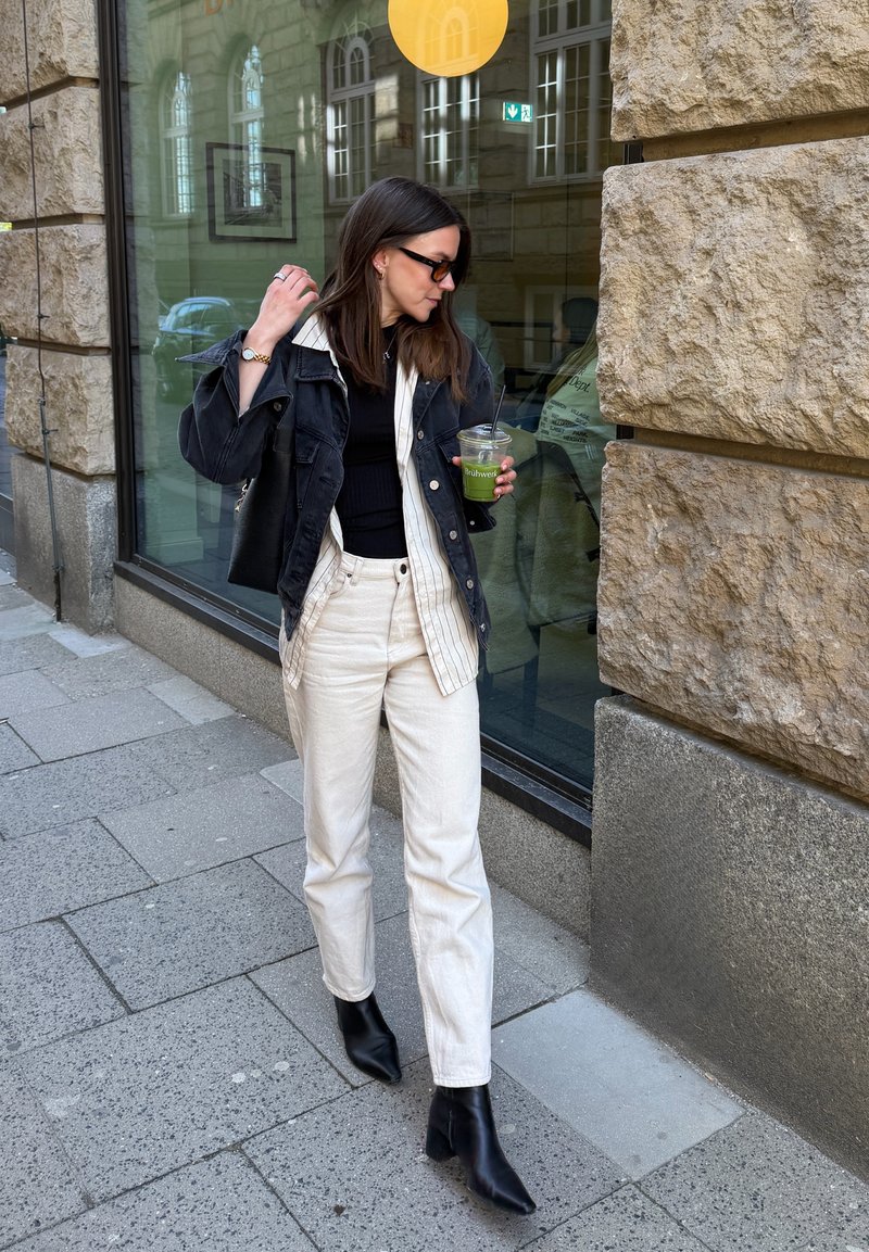 Femme en veste noire, chemise rayée, pantalon écru et bottes noires marchant sur le trottoir de la ville en tenant une boisson verte dans un gobelet en plastique avec paille.