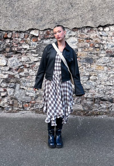 Chaqueta de cuero sintético negra sobre un vestido de cuadros con volantes, combinada con botas negras hasta la rodilla y un bolso de cruzar. Fondo de pared de piedra.