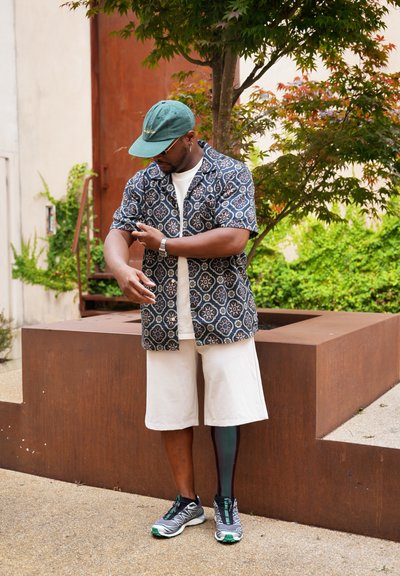 Camisa de manga corta con estampado en azul marino oscuro con motivos florales, camiseta blanca, pantalones cortos beige y zapatillas grises con acentos verdes y una pierna prostética.