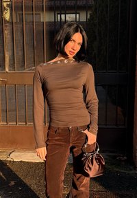 Bluză maro cu mâneci lungi, cu textură adunată și nasturi de accent argintii pe umăr, asortată cu pantaloni maro din catifea de tip cord și o geantă mică maro.