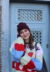 Jeune femme aux longs cheveux portant un bonnet bordeaux et un pull coloré à motif fraise, souriant devant une porte décorative bleue.