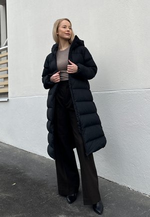 Abrigo puffer negro con capucha, con un diseño largo y acolchado. La prenda tiene una textura brillante y se combina con pantalones oscuros y botas.