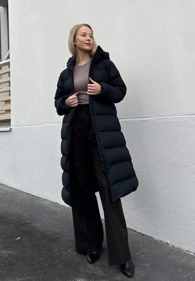 Abrigo puffer negro con capucha, con un diseño largo y acolchado. La prenda tiene una textura brillante y se combina con pantalones oscuros y botas.