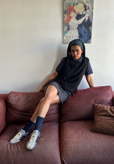 Persona sentada en un sofá marrón, vestida con una blusa sin mangas oscura sobre una camisa azul, una falda de mezclilla y zapatillas deportivas plateadas con calcetines oscuros.