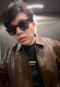 Personne portant de grandes lunettes de soleil foncées, une veste en cuir brun foncé et une chemise noire avec un texte blanc dans un environnement intérieur.