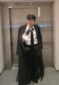 Personne portant un long manteau noir, une chemise blanche, une cravate noire et un pantalon à fines rayures. Des gants noirs et des lunettes complètent la tenue. Fond d'ascenseur.