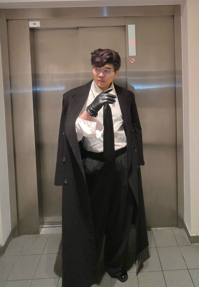 Personne portant un long manteau noir, une chemise blanche, une cravate noire et un pantalon à fines rayures. Des gants noirs et des lunettes complètent la tenue. Fond d'ascenseur.