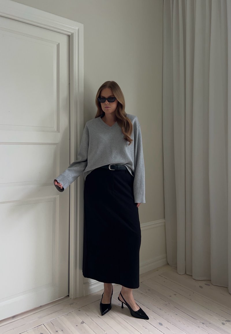 Femme portant des lunettes de soleil, un pull gris, une jupe midi noire et des talons noirs, debout à l'intérieur près d'une porte blanche et d'un rideau.