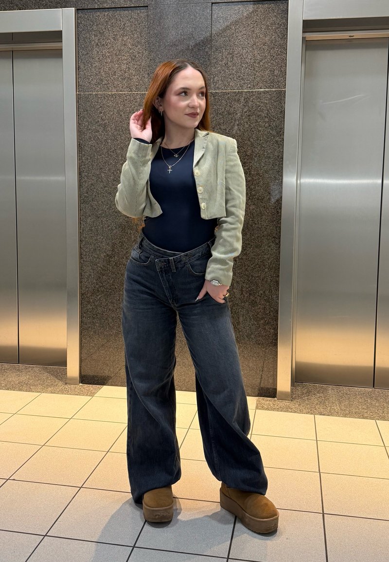 Jeune femme debout entre deux ascenseurs en acier inoxydable, portant une veste verte courte, un haut noir, un jean large et des chaussures compensées marron.