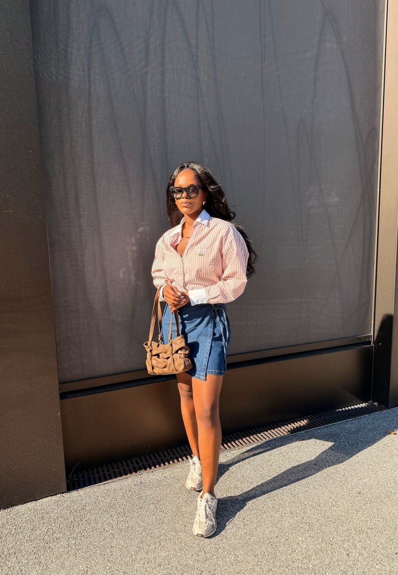 Donna con occhiali da sole, camicia rosa a righe, gonna di jeans, sneakers bianche, tiene una borsa marrone, in piedi alla luce del sole contro un muro scuro.