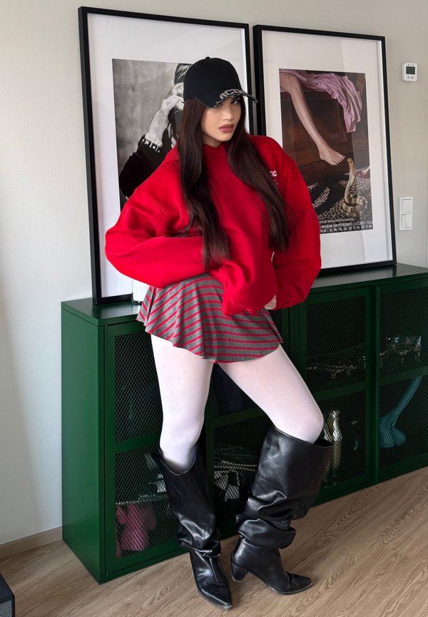 Fiatal nő pózol bent, piros kapucnis pulóvert, csíkos szoknyát, fehér harisnyát, fekete térdig érő csizmát és fekete sapkát viselve, mögötte keretezett műalkotással.