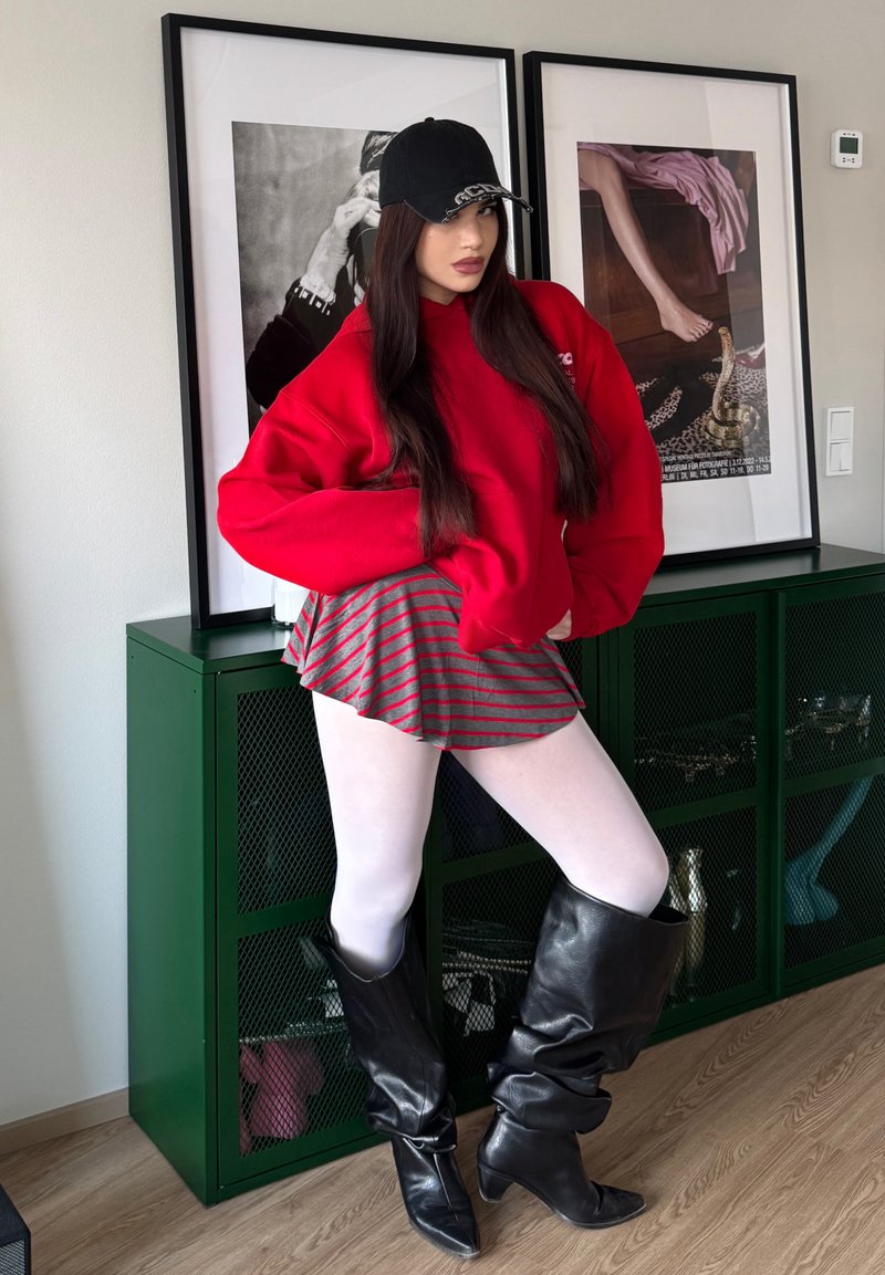 Νεαρή γυναίκα ποζάρει μέσα, φορώντας κόκκινο φούτερ με κουκούλα, ριγέ φούστα, λευκές καλσόν, μαύρες μπότες έως το γόνατο και μαύρο καπέλο, με κορνιζαρισμένη τέχνη στο βάθος.