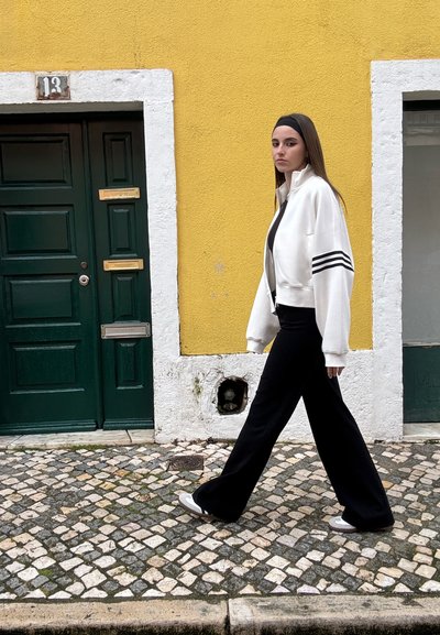 Chaqueta bombers blanca con rayas negras, pantalones anchos negros y zapatos grises, frente a una pared amarilla y una puerta verde.