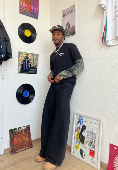 Jeans anchos negros, combinados con una camisa de manga larga de teñido anudado. Los accesorios incluyen un collar de cadena y una gorra estampada. Discos de vinilo en el fondo.
