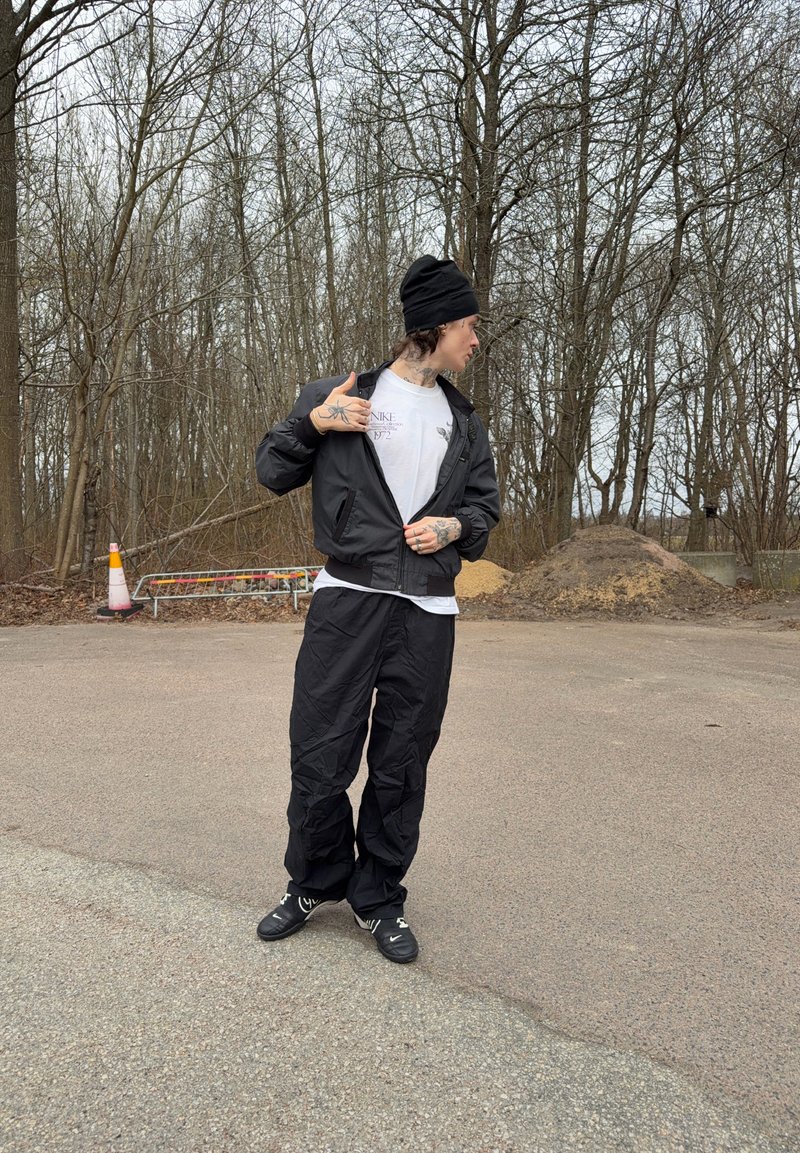 Personne portant un bonnet noir, une veste noire ouverte laissant apparaître un T-shirt blanc Nike, un pantalon noir et des chaussures noires, debout sur une zone pavée près d'arbres sans feuilles.