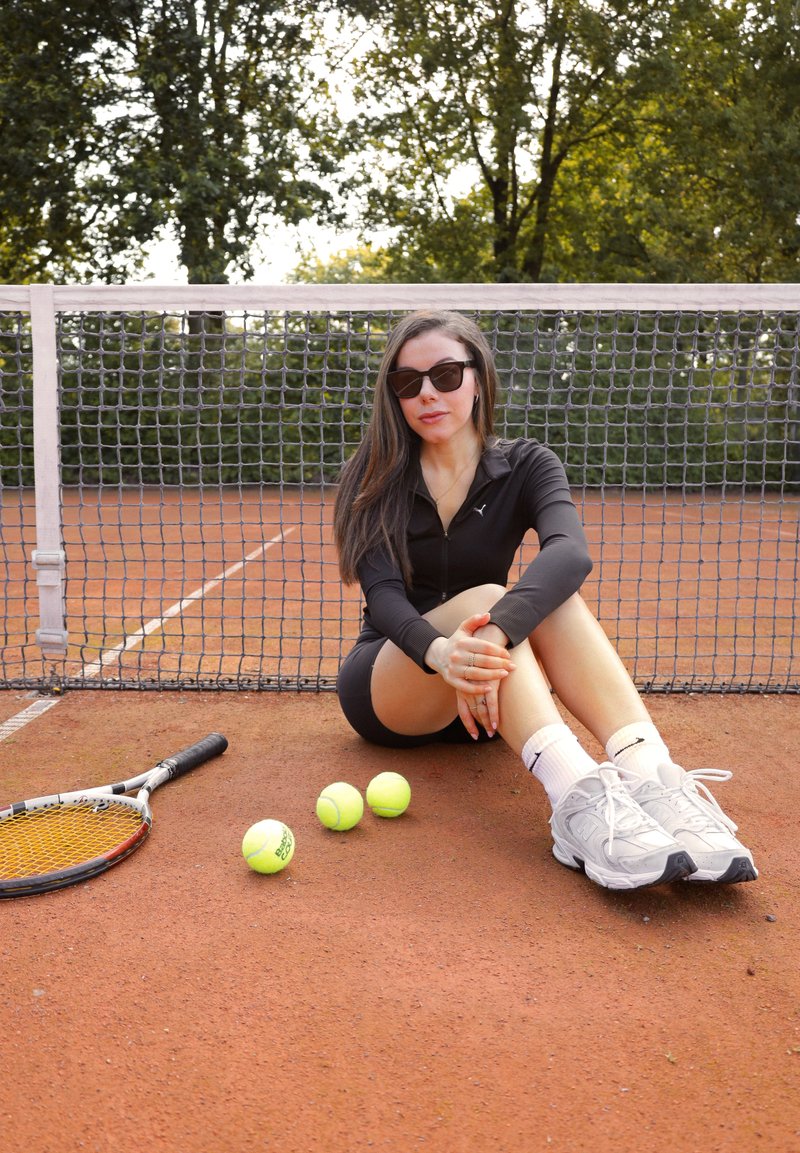 Μαύρο αθλητικό φερμουάρ μπουφάν, εφαρμοστά μαύρα σορτσάκια, γκρι αθλητικά παπούτσια, καθισμένος σε γήινο γήπεδο τένις με τρεις κίτρινες μπάλες τένις και μια ρακέτα κοντά.
