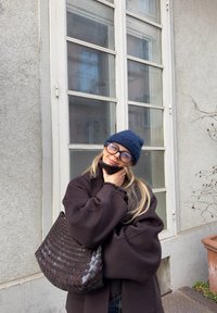 Femme aux longs cheveux blonds portant un bonnet bleu marine, de grandes lunettes et un manteau foncé, posant avec la main sur le menton et tenant un sac tissé marron foncé.