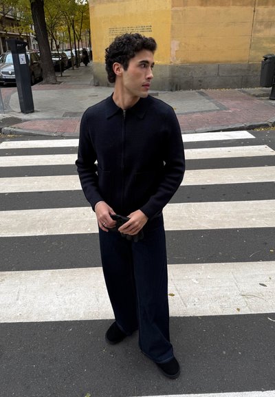 Joven con chaqueta y pantalones negros sosteniendo guantes, de pie en un paso de peatones junto a un edificio beige y coches aparcados en una calle de la ciudad.