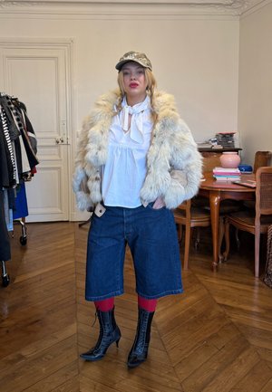 Chaqueta de piel sintética en tonos crema y marrón claro, blusa blanca con detalle de lazo, culottes de mezclilla oscuros, medias rojas y botas de tacón de encaje negras.