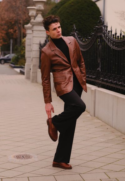 Blazer de cuero marrón sobre un cuello de tortuga negro, combinado con pantalones negros y zapatos de vestir marrones, posando sobre una acera con patrón y vegetación.