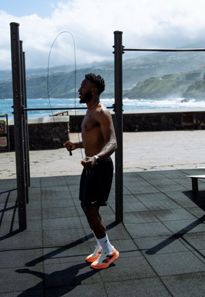Atleta che salta la corda in una palestra all'aperto; indossa pantaloni corti neri e scarpe da corsa arancioni. Lo sfondo presenta l'oceano e un paesaggio collinare.