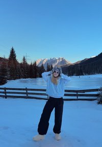 Jeune femme en vêtements d'hiver debout dans un champ enneigé avec une clôture en bois, des pins et une chaîne de montagnes éclairée par le soleil sous un ciel bleu clair.