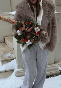 Manteau en fausse fourrure marron, sweat à capuche blanc zippé, pantalon de survêtement gris. Tenant une couronne avec des éléments de pin, du coton, des accents rouges et des bâtons de cannelle, debout sur la neige.