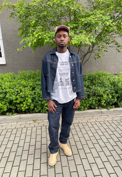 Chaqueta de mezclilla sobre una camiseta gráfica blanca, combinada con jeans oscuros y botas de tobillo beige. El entorno cuenta con arbustos verdes y una zona pavimentada.