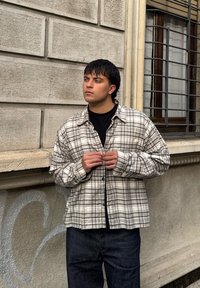 Camisa aos quadrados em bege e cinza, corte folgado, abotoada à frente, usada sobre uma t-shirt preta; combinada com jeans escuros. Fundo texturizado.