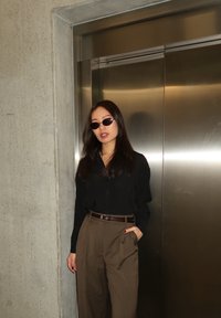 Femme portant des lunettes de soleil, une chemise noire et un pantalon marron, debout avec une main dans la poche à côté d'une porte d'ascenseur en acier inoxydable fermée.