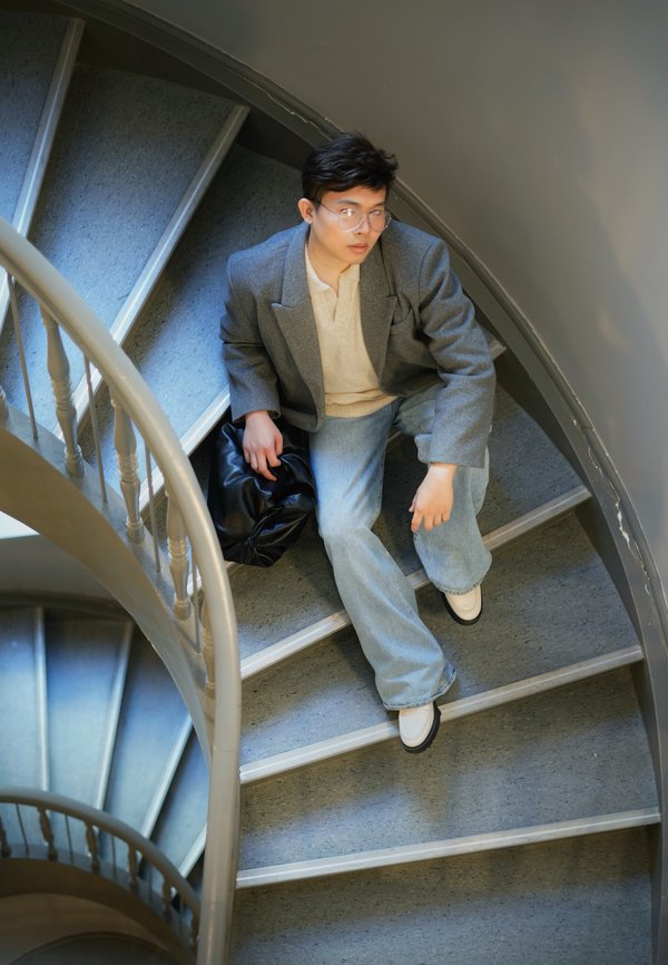 Persona seduta su una scala a chiocciola grigia, indossa un blazer grigio, un maglione beige e jeans a zampa d'elefante blu. Presenza di una borsa nera e scarpe bianche.